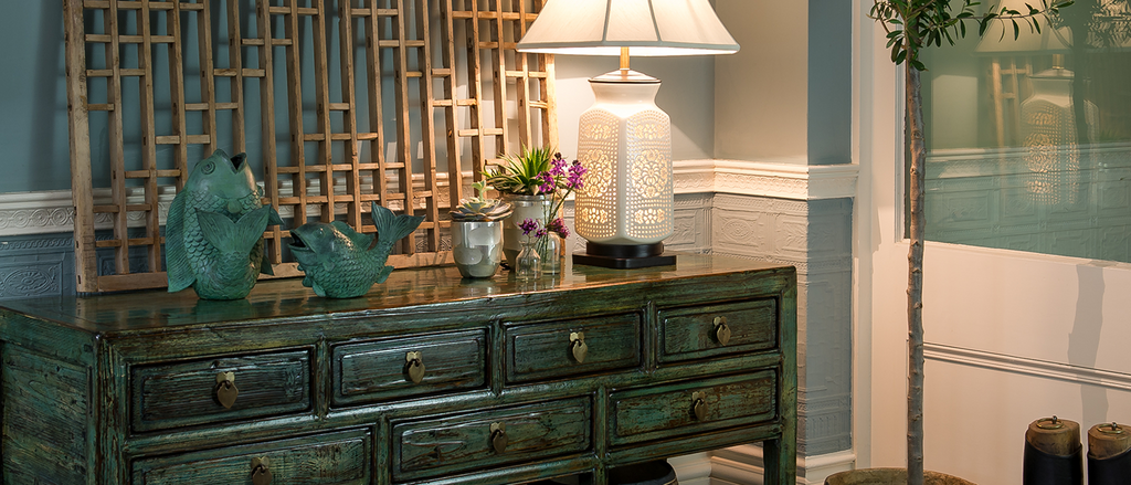 orchid eclectic interior with oriental accents. green distressed lacquered dresser styled with traditional Chinese window panel, white lace ceramic table lamp and two metal fish statues 