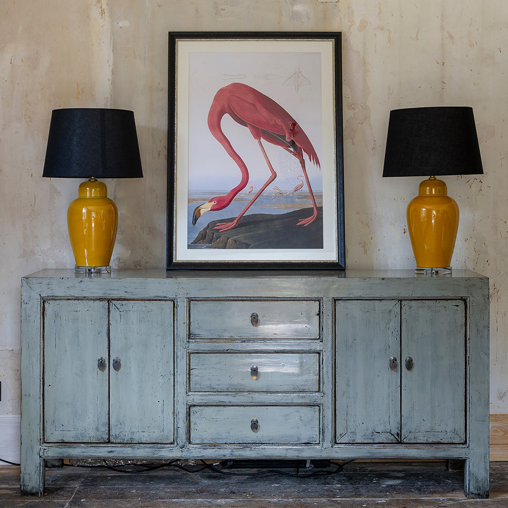 distressed grey sideboard with two cabinets and three drawers in the middle. styled with ceramic mustard lamps and vintage pink flamingo print