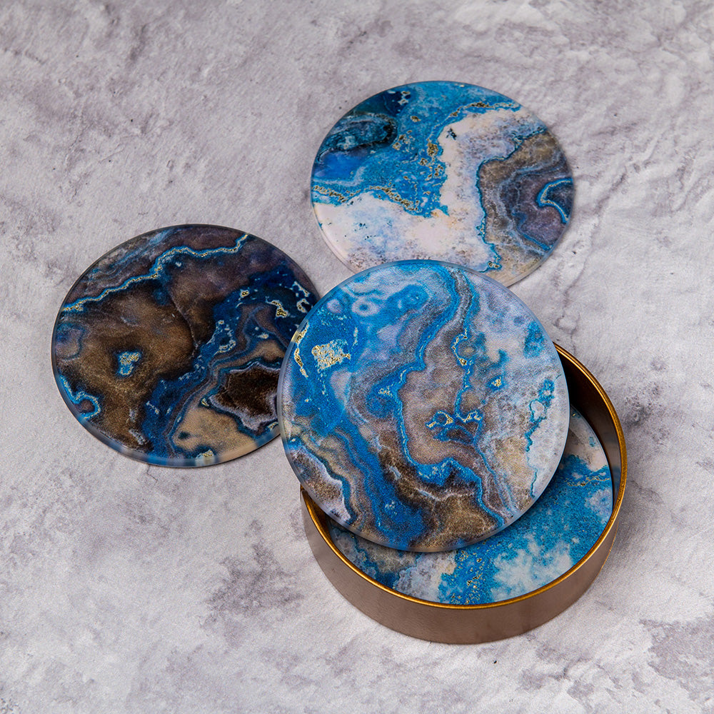 A set of 4 coasters presented in a gold storage container. Each coaster has a cork backing and is finished with a blue marble effect glass topping. 