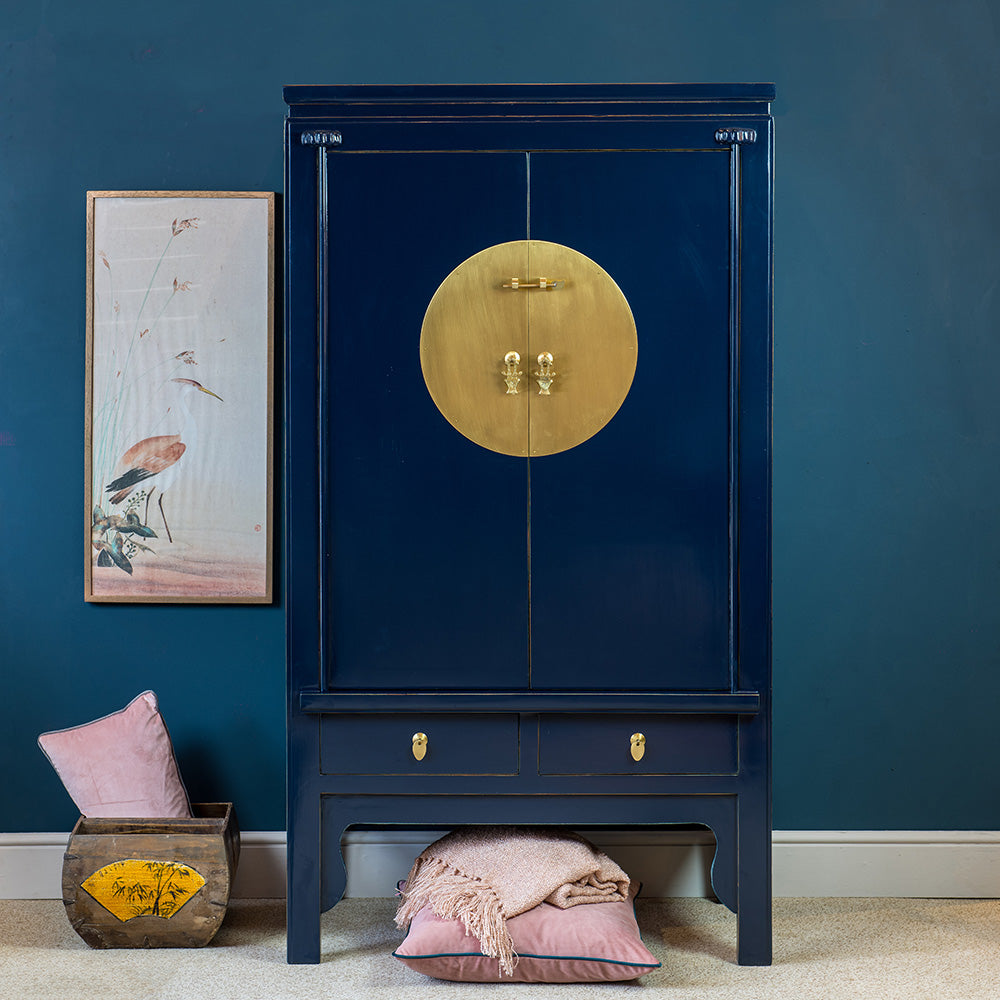 A dark blue wedding wardrobe featuring a gold brass circular panel and traditional Chinese hardware. It has a double door cabinet and two drawers. 